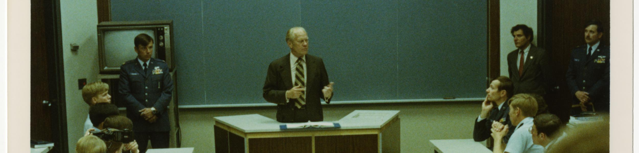 President Ford stands behind a desk with an open binder, talking to a room full of military members.