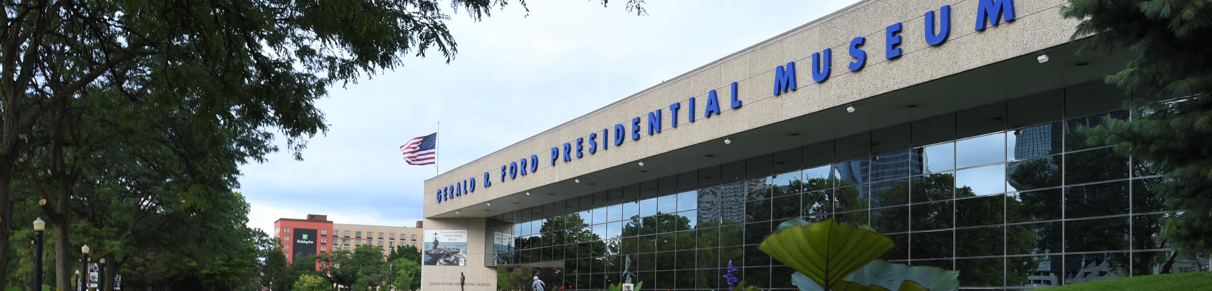 The exterior of the museum showcases gardens filled with colorful flowers, green lawns and the building has large blue block letters reading "Gerald R Ford Presidential Museum"