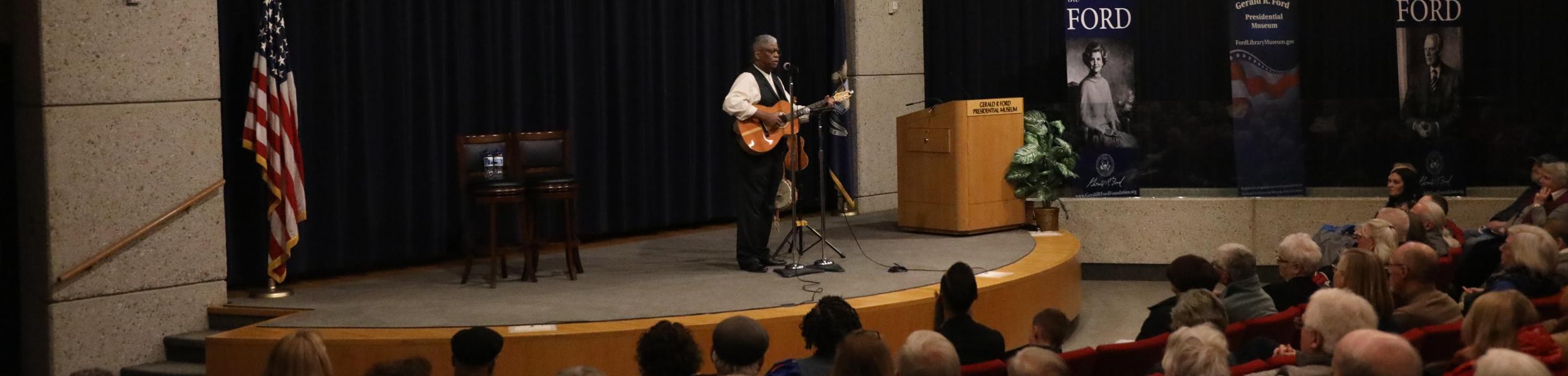 Reverend Jones stands alone on a stage, playing a guitar as the audience looks on.