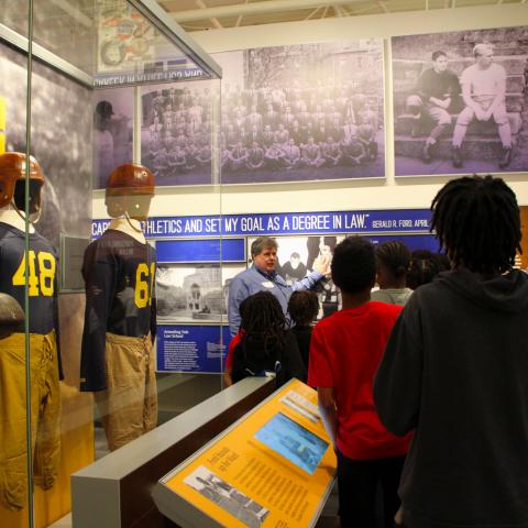 A museum staff member leads a group of students and adults through an exhibit.