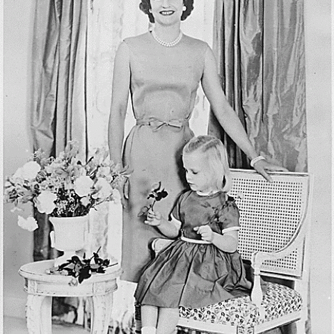 Betty Ford stands while a young Susan Ford is seated on a small chair in front of her. This photo was created in 1961