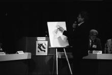 artist standing at an easel speaking into a microphone while drawing