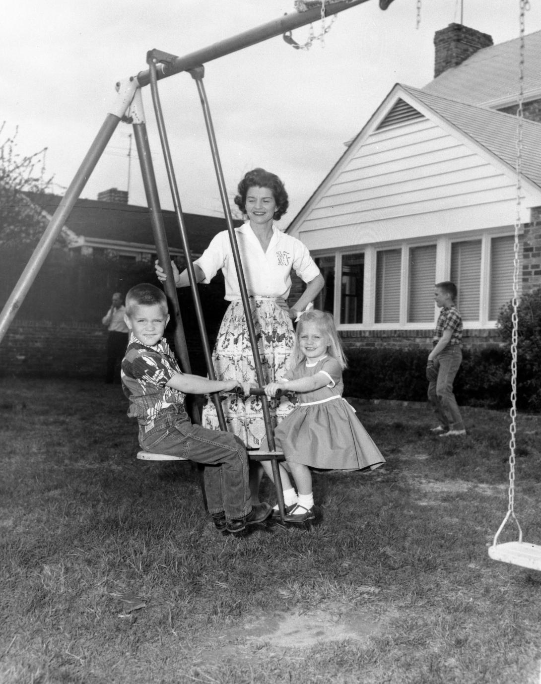 H0009-3. Betty watches as her children Steve and Susan ride on a glider in the back yard of their home at 514 Crown View Drive, Alexandria, Virginia. Her son Michael appears in the background. 1962.