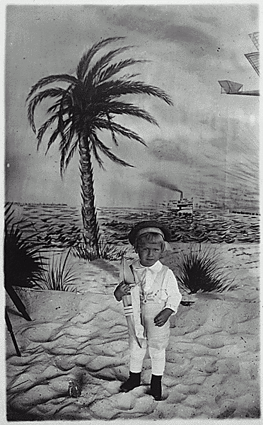 H0016-4 and H0020-2. Gerald R. Ford, Jr. holding a toy boat, poses for portrait; wearing sailor cap, standing on sand, painted backdrop of palm tree, steamship, and airplane in flight, in background. 1916.